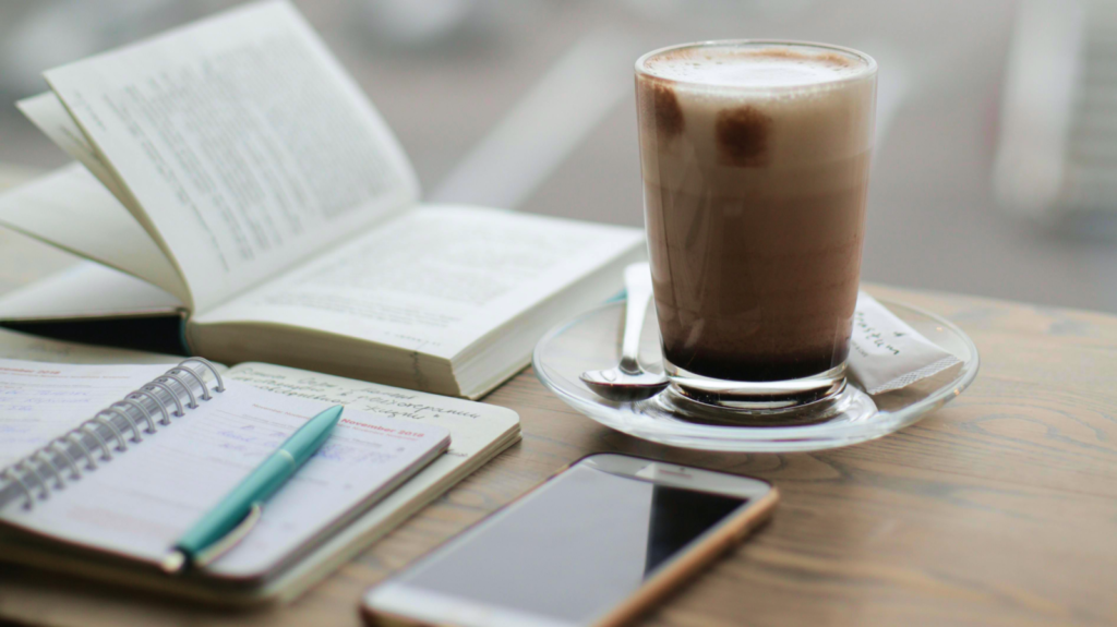 Diabetes Selbstmanagement. Auf einem Holztisch liegt ein Buch zur Information sowie ein Diabetes-Tagebuch mit Kugelschreiber, ein Smartphone und ein Kaffee. 